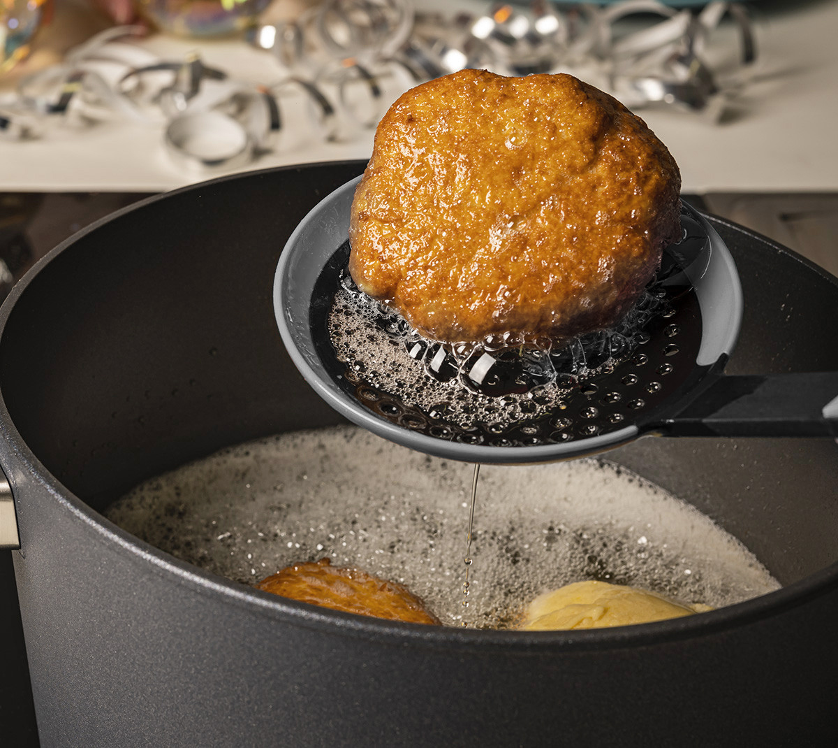 Cook it Schaumlöffel Mit einer grauen Cock it Schöpfkelle wird ein ausbackener Fastnachtkrapfen aus dem Topf entnommen und über dem Topf abgetropft.