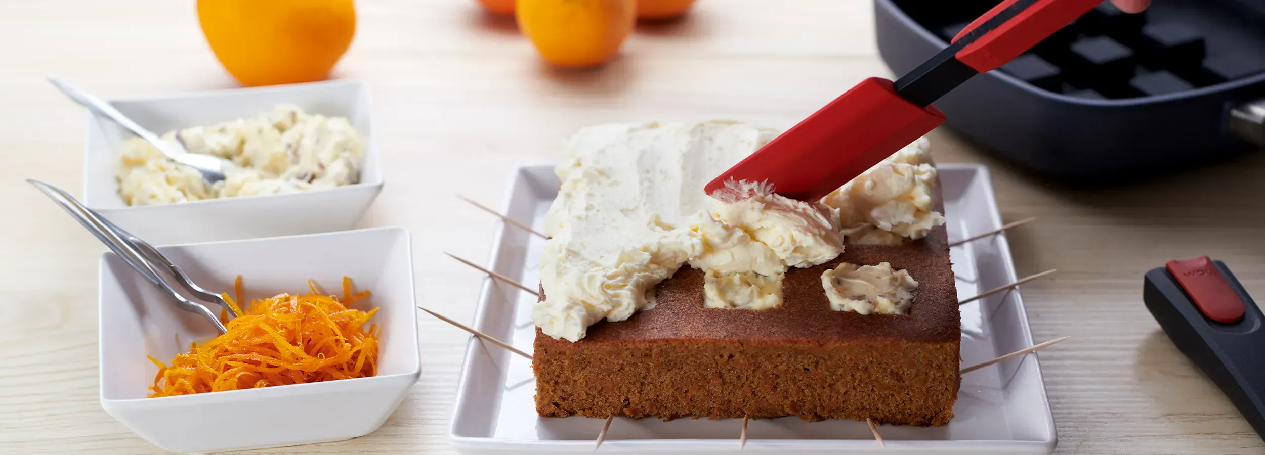 Mit dem Teigschaber von Cock it wird die Creme auf dem frisch gebackenen Kuchen verteilt. Daneben liegt eine Pfanne mit Silikon-Backeinsatz und abgenommenem Stiel sowie weitere Toppings und Orangen.