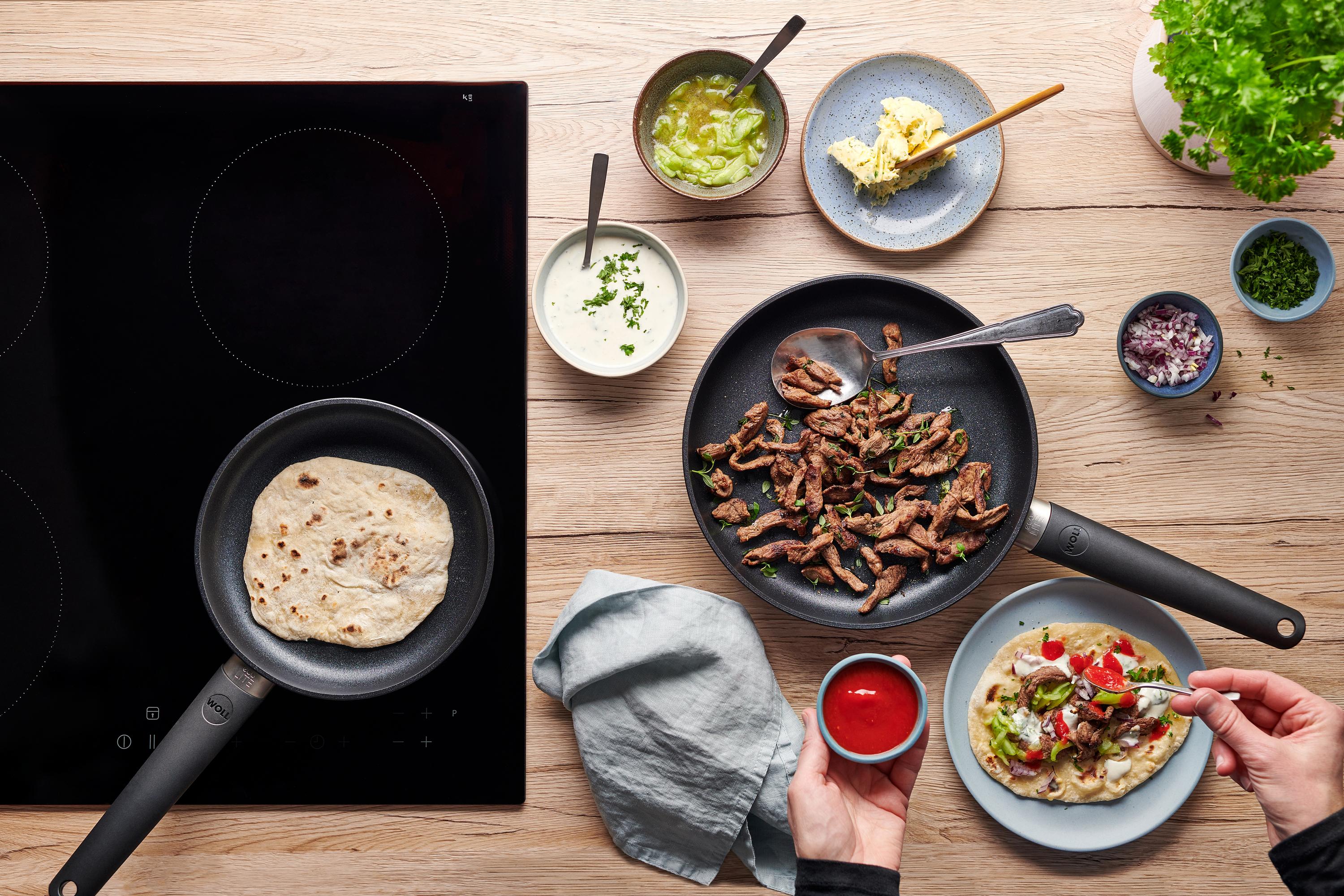 Eco Lite QXR Flachpfanne In der runden Eco-Lite-QXR-Pfanne werden Fladenbrote gebacken. Daneben liegt in einer Pfanne angebratenes Lamm und auf einem Teller ist Döner zubereitet.