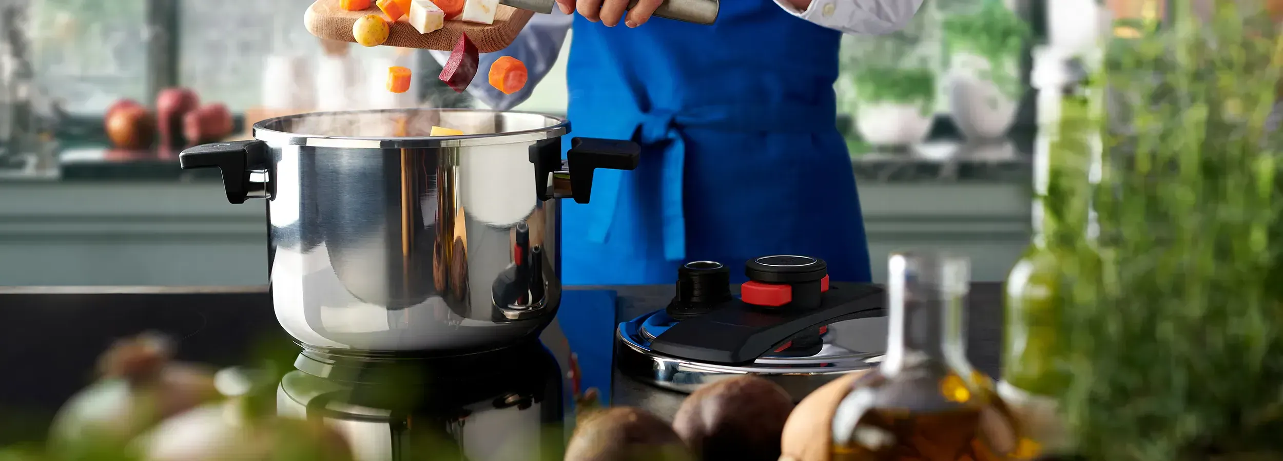 Auf dem Herd steht ein dampfender Pressure-Pro-Topf, während das frisch geschnittene Gemüse mit dem Messer vom Brett in den Topf gegeben wird. Der Deckel liegt rechts auf der Arbeitsplatte.
