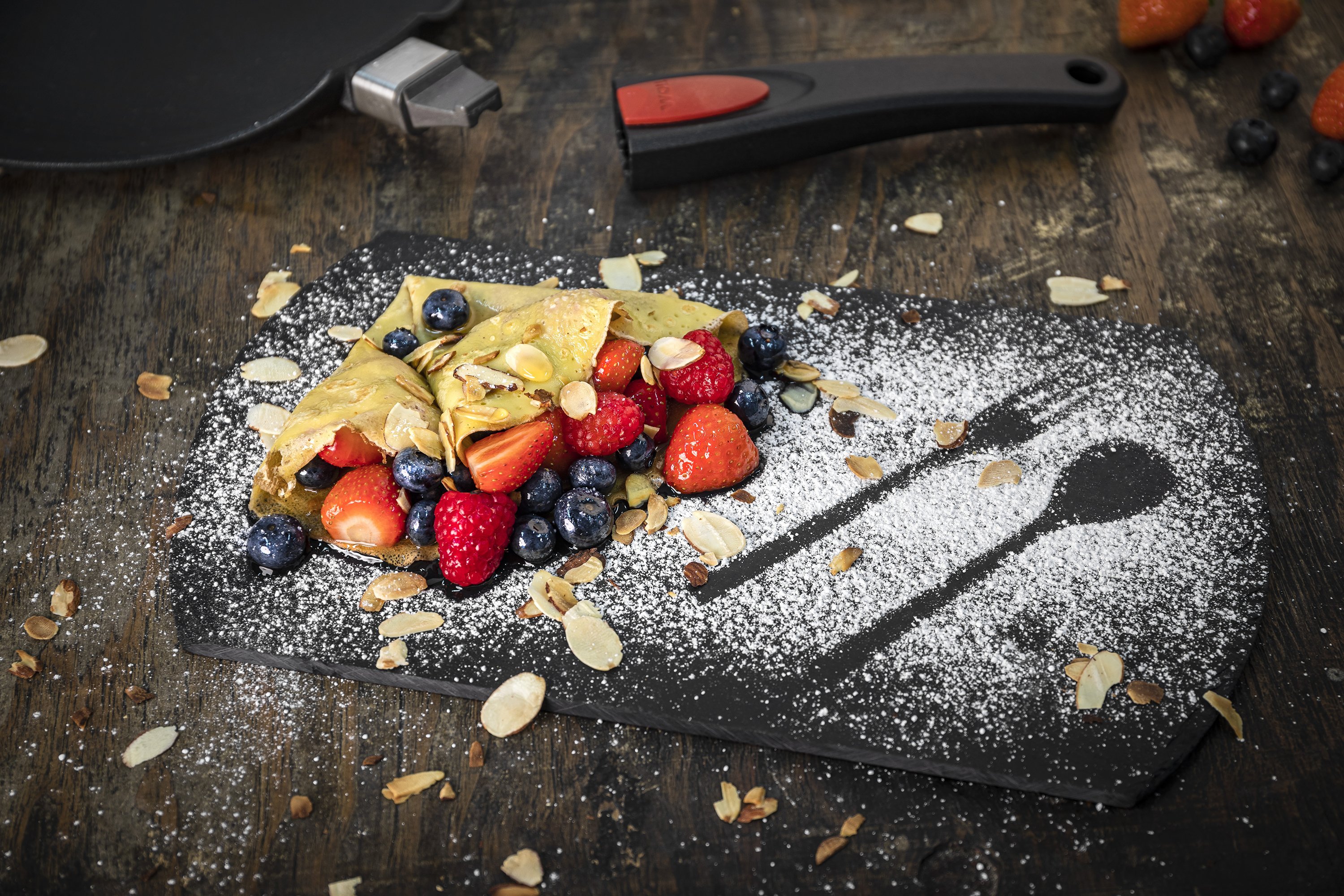 Auf einer Platte liegt Crêpe mit marinierten Früchten, dieser wurde mit Puderzucker und Mantelblättchen garniert. Im Hintergrund sieht man eine runde Crêpepfanne aus der Serie Titan Best mit abgenommenem Stiel.