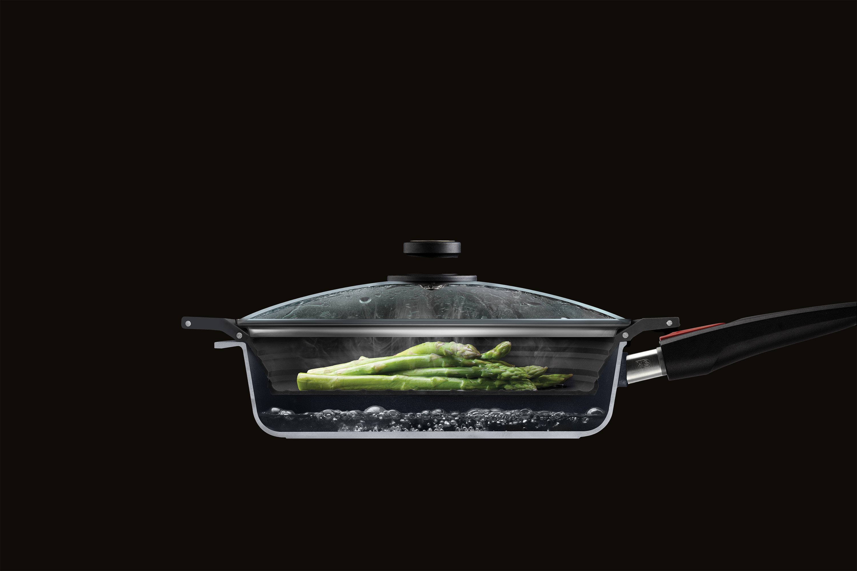 Im Querschnitt ist eine halbierte Pfanne mit Glasdeckel zu sehen, in der grüner Spargel mit dem Steam it multifunktionalen Silikoneinsatz gedämpft wird. 