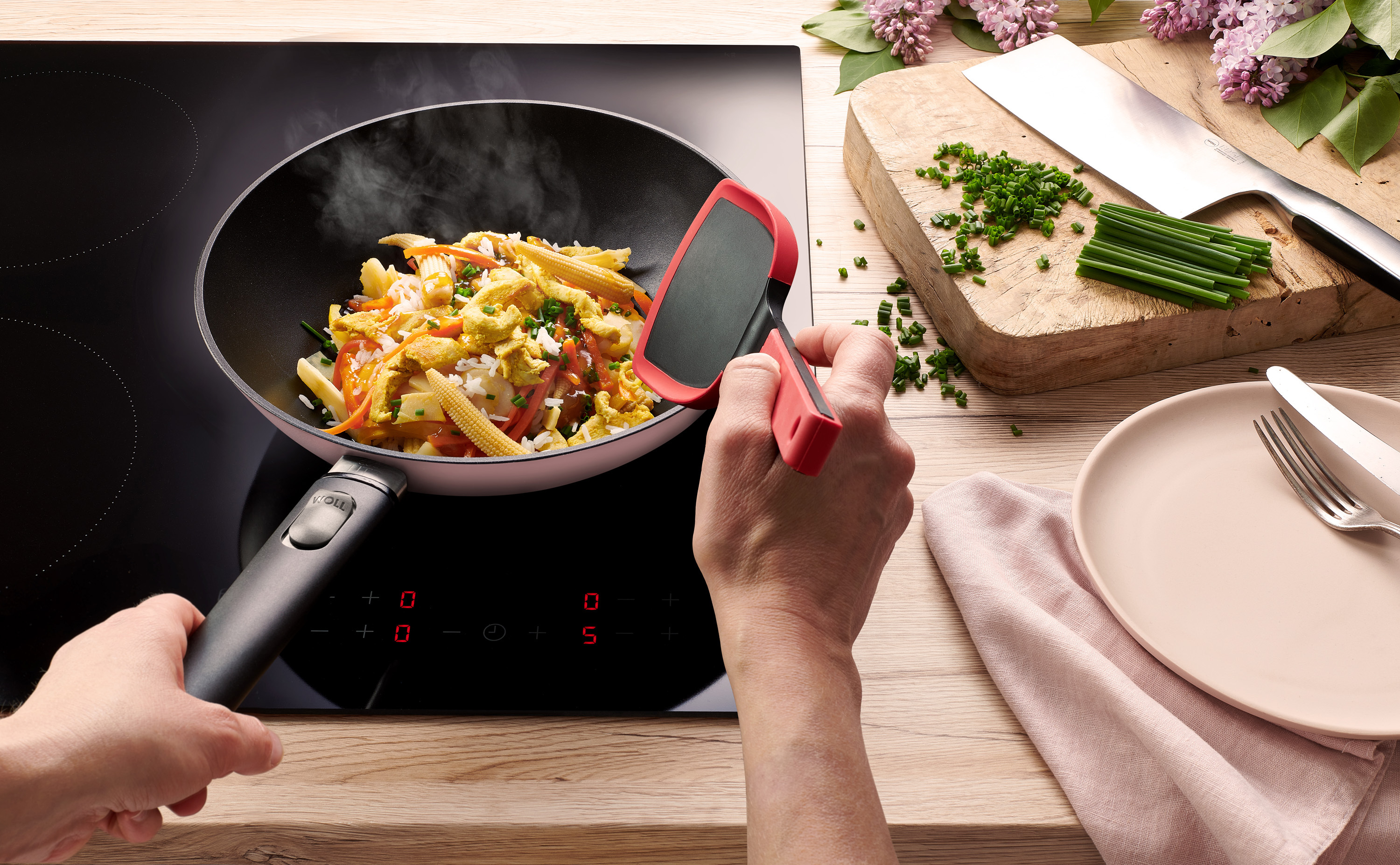 Ecolour Wok mit Deckel, Ø 24 cm Eine runde, hellrosa Ecolour Wokpfanne steht auf dem Herd, mit einem Cock it wird ein dampfendes Gericht gewendet. Das Edge Chinesisches Kochmesser liegt daneben.